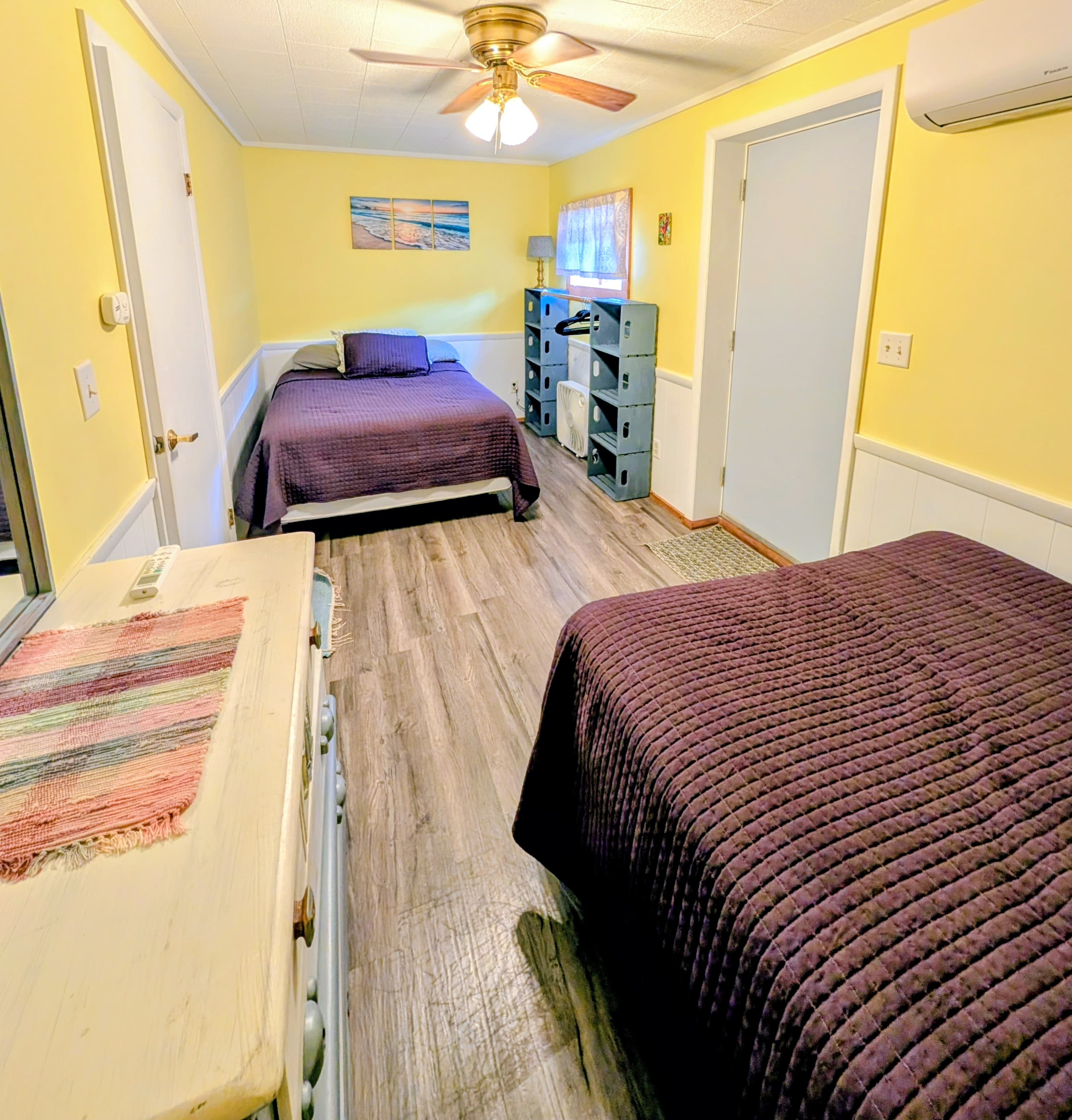 A cozy bedroom featuring two beds with purple bedding, a dresser, and yellow walls.