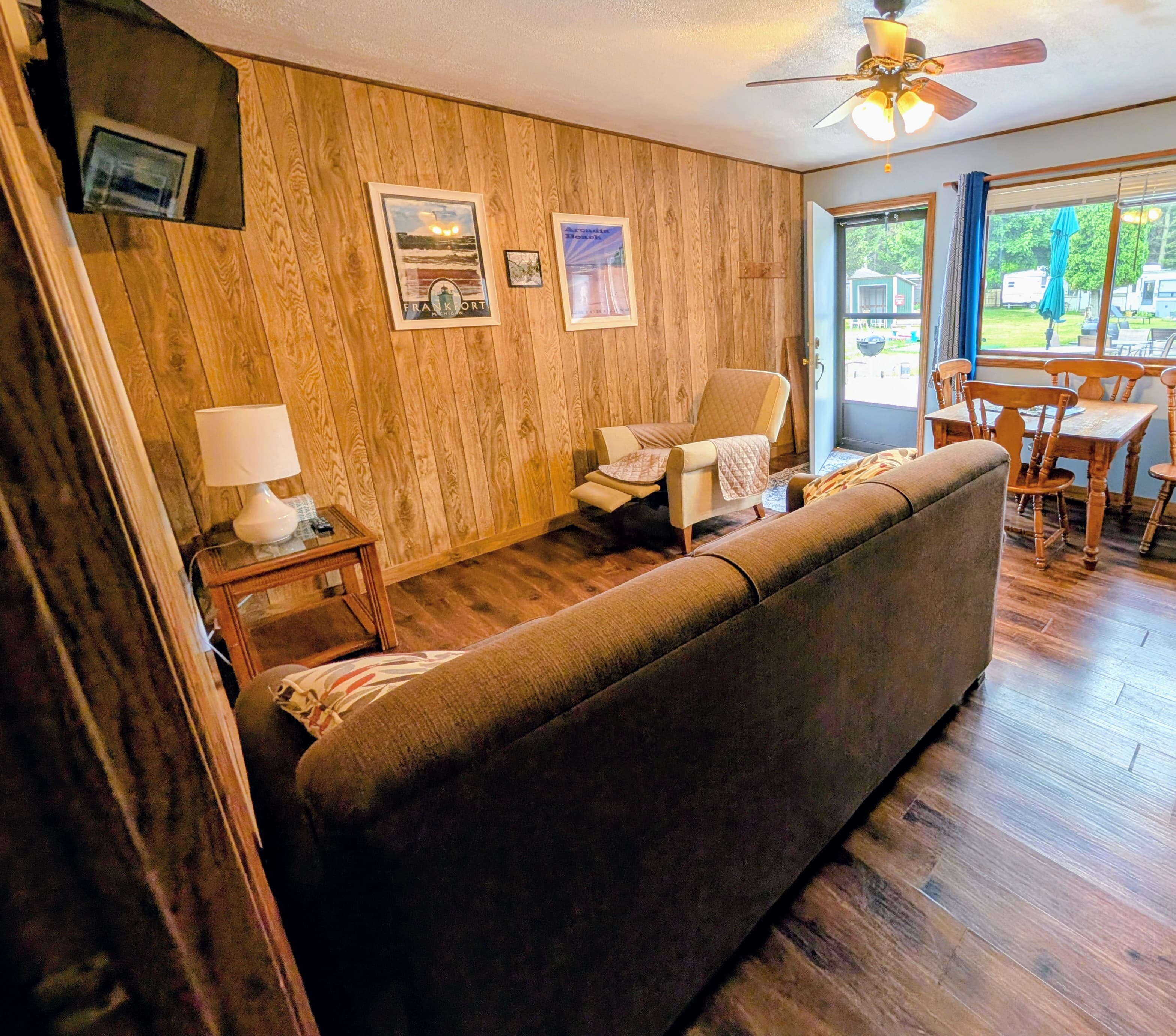 A cozy living room featuring a brown sofa, a lamp, wooden walls, and a dining area with a view of the outdoors.