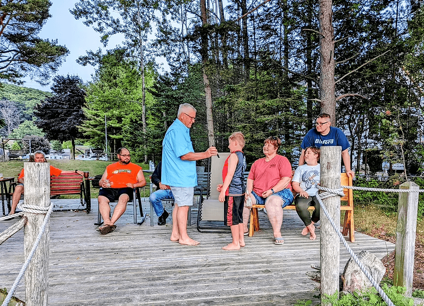 A group of people gathers by a lakeside deck, with an adult speaking to a child while others watch.