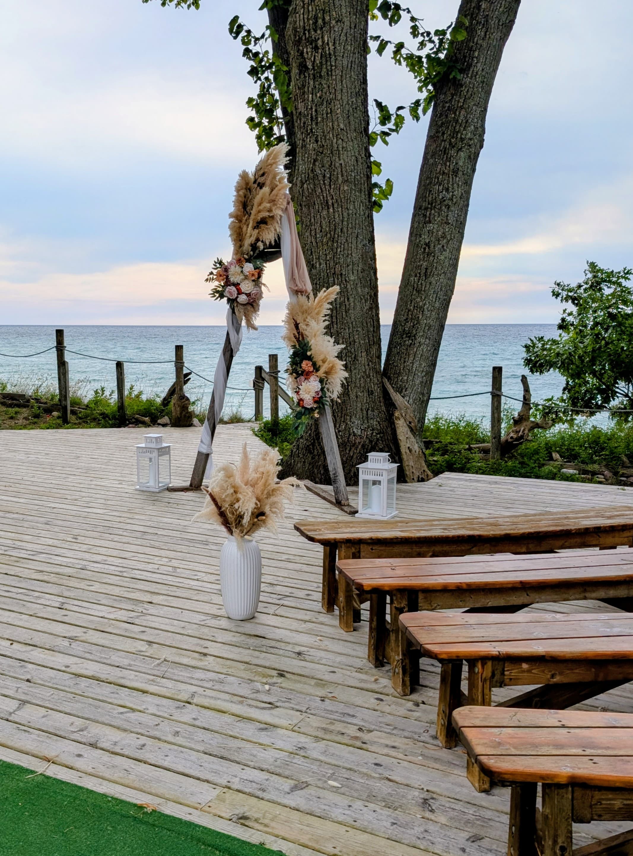 A decorated wedding arch stands on a wooden platform by the seaside, surrounded by rustic benches and lanterns.