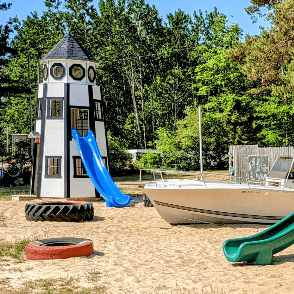 A colorful play structure resembling a lighthouse with slides, surrounded by sand and a boat.