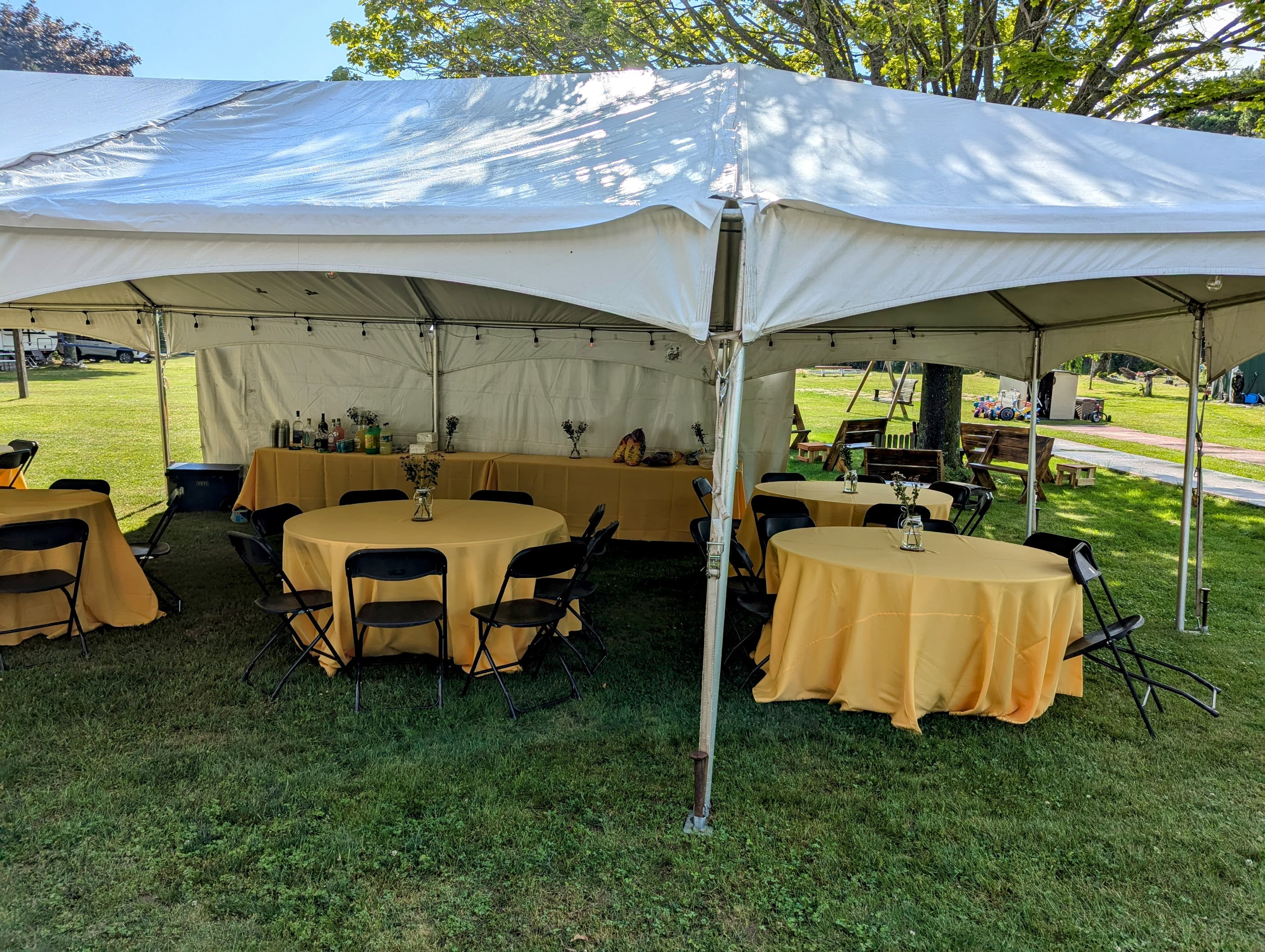 A large outdoor tent with yellow tablecloths and black chairs set up on a grassy field.