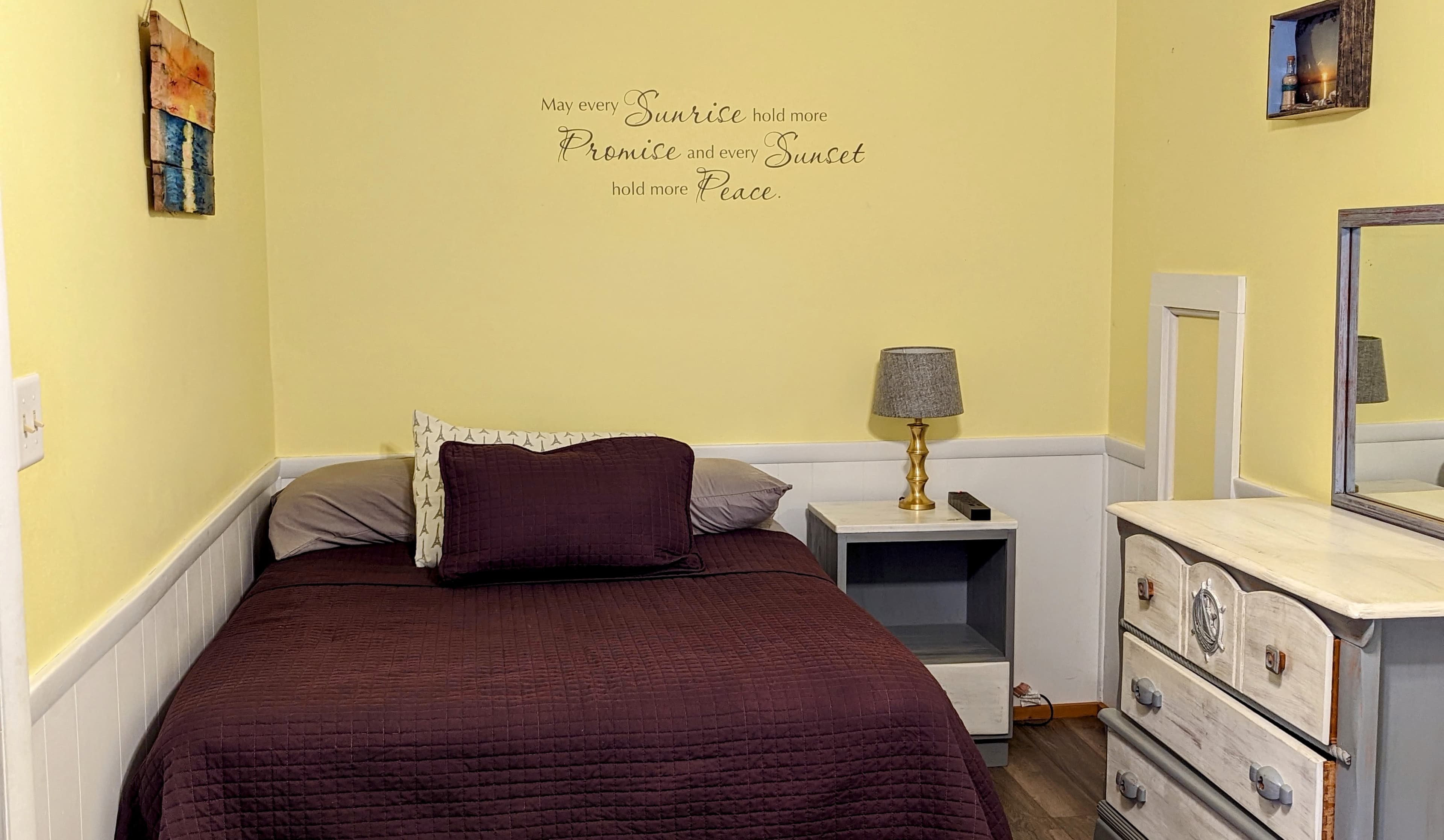 A cozy bedroom featuring a bed with a maroon quilt, a bedside lamp, and a mirrored dresser against a yellow wall with an inspirational quote.