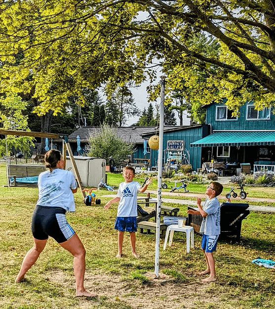A woman plays a game with two boys outdoors under a large tree, featuring a ball and a scoring pole in the backyard.