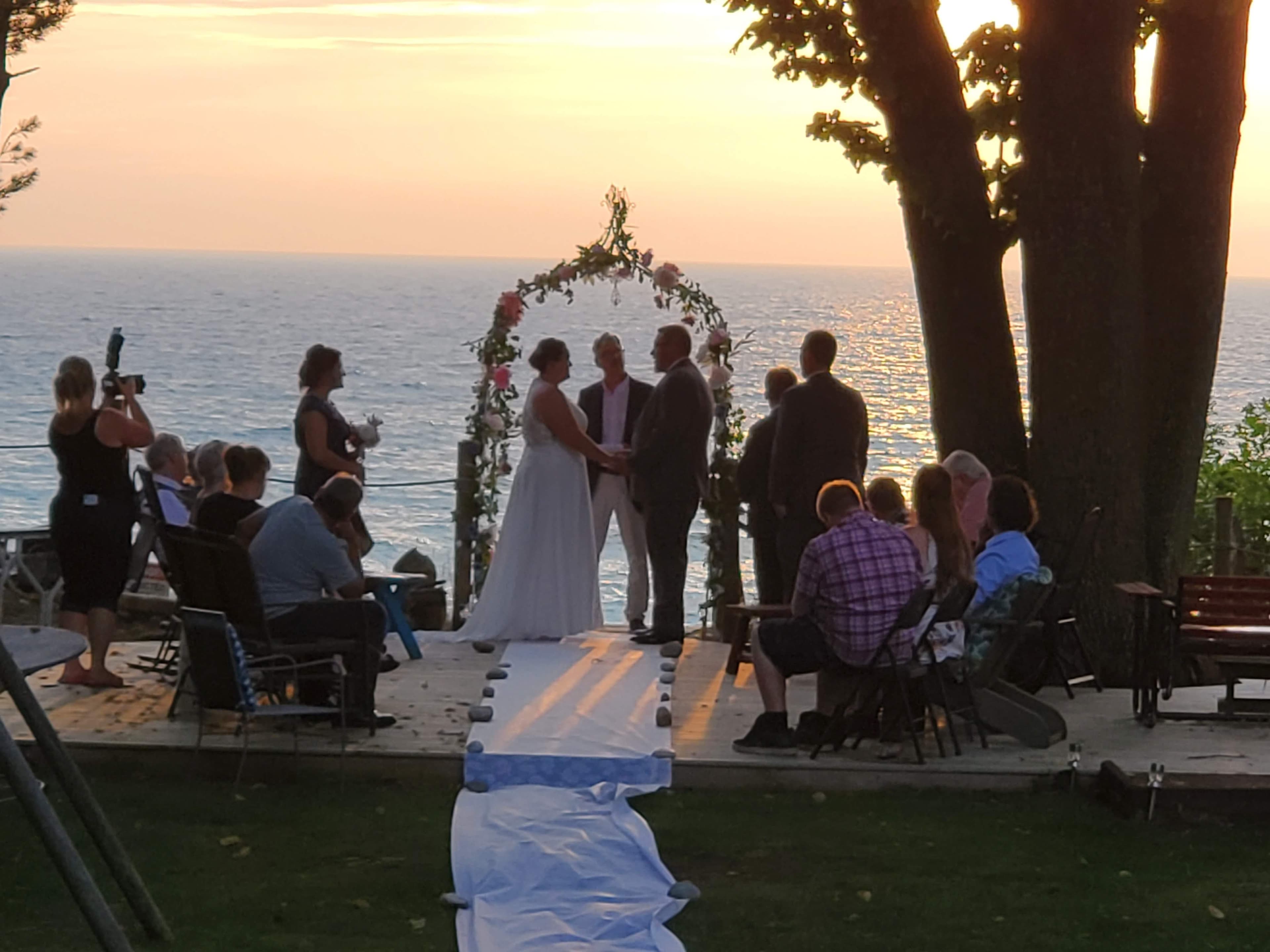 A couple exchanges vows at a sunset wedding ceremony by the water, surrounded by guests.