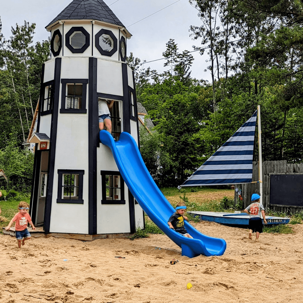 Two children play on a sandy playground with a lighthouse slide, while another climbs inside.