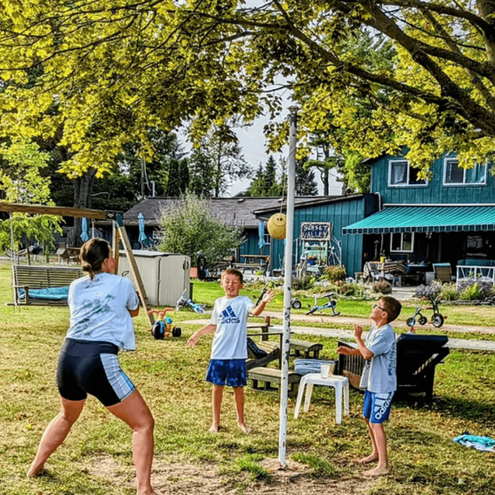 A woman plays an outdoor game with two children near a house surrounded by trees.