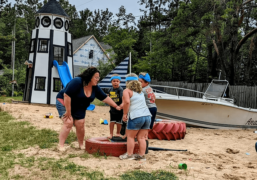 A group of children gather around a woman on a sandy beach near a playground structure and a boat.