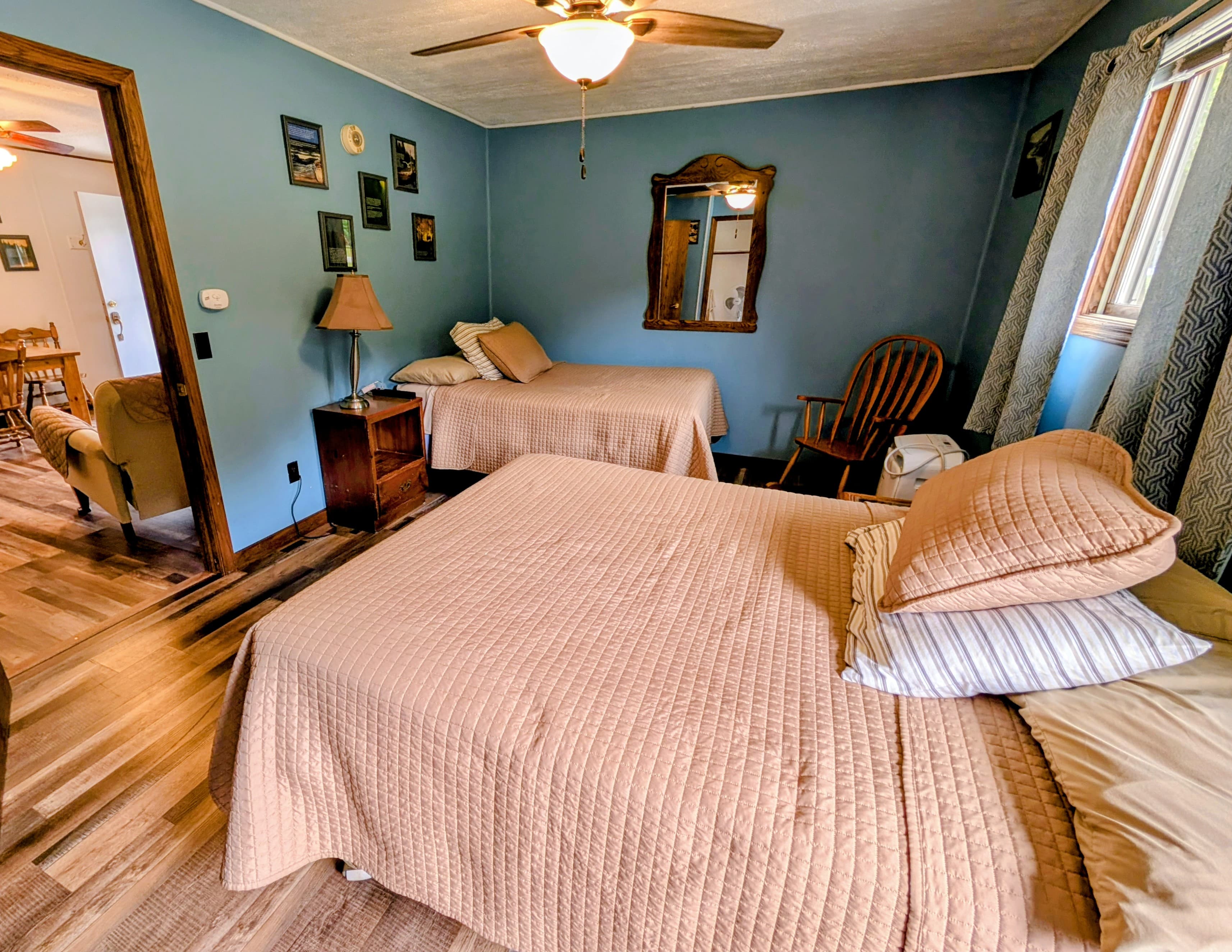 A cozy bedroom featuring two beds with beige quilted covers, a lamp, a mirror, and wooden furniture against a blue wall.