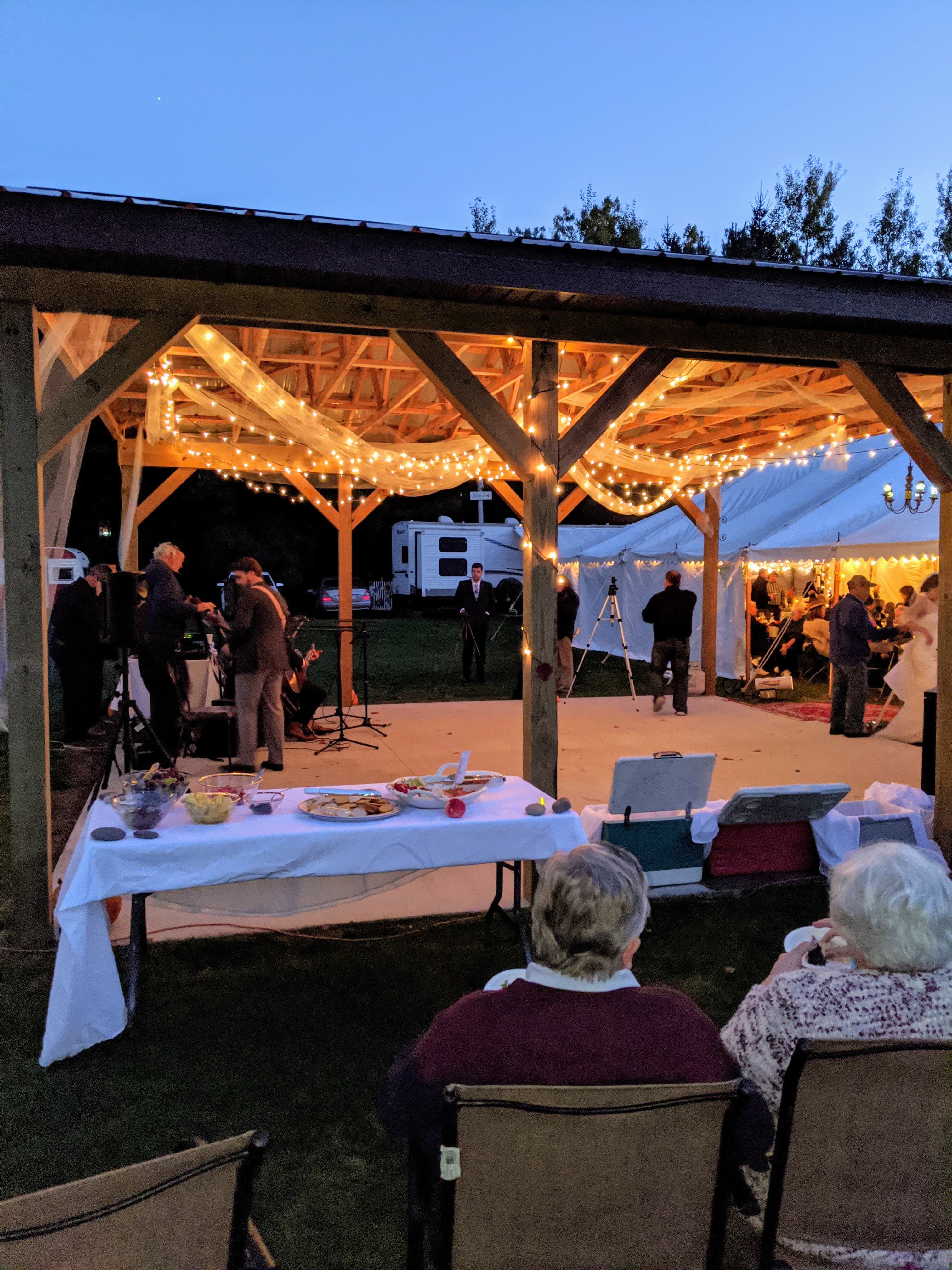 An outdoor gathering under a lit pavilion with guests enjoying food while a band plays nearby.