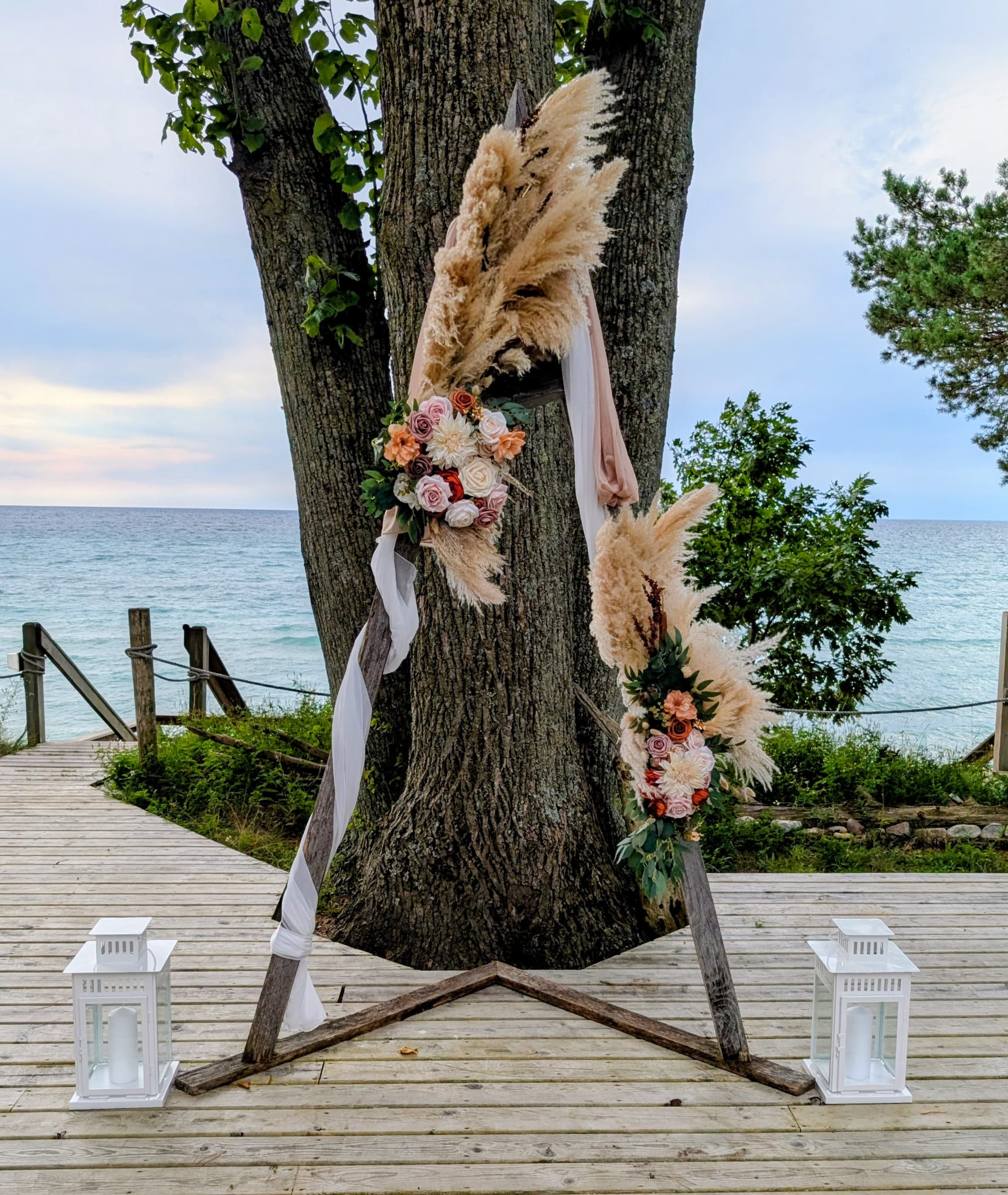 An elegant wedding arch adorned with pampas grass and flowers, set against a serene waterfront backdrop.
