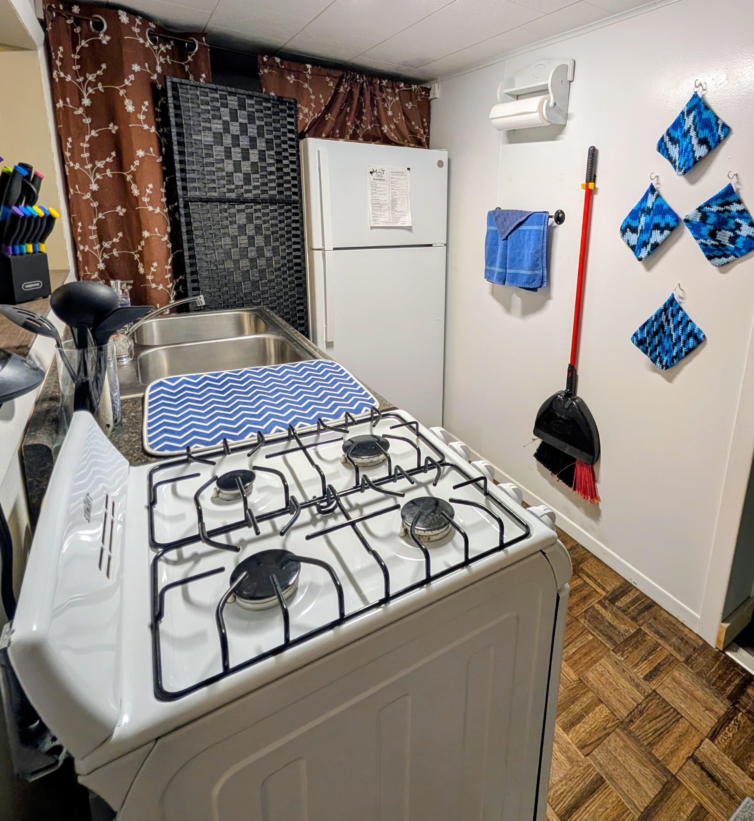 A compact kitchen featuring a gas stove, sink, refrigerator, and colorful cleaning supplies.