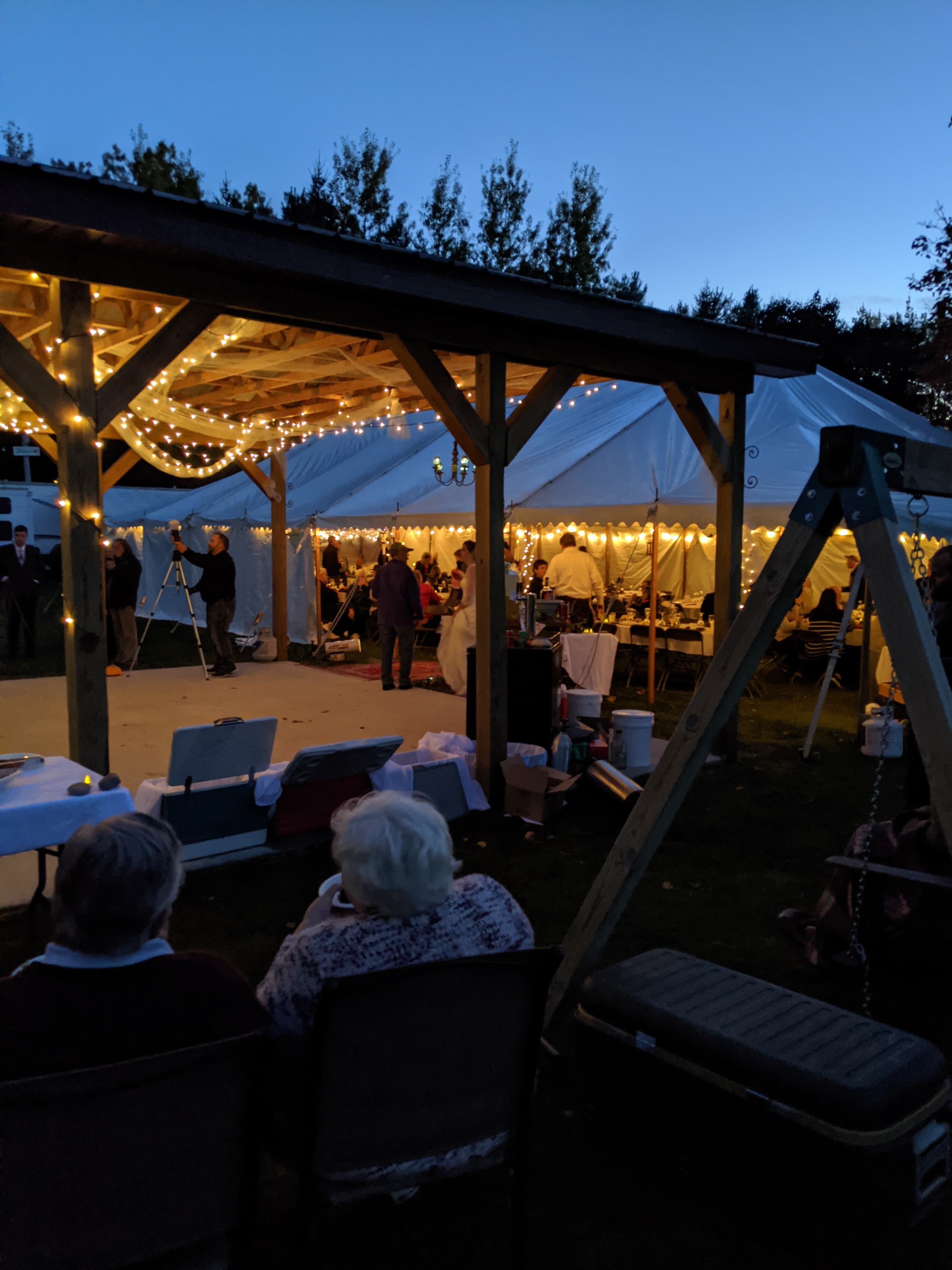 A lively outdoor event under twinkling lights with guests gathered in a tent and two seated individuals in the foreground.