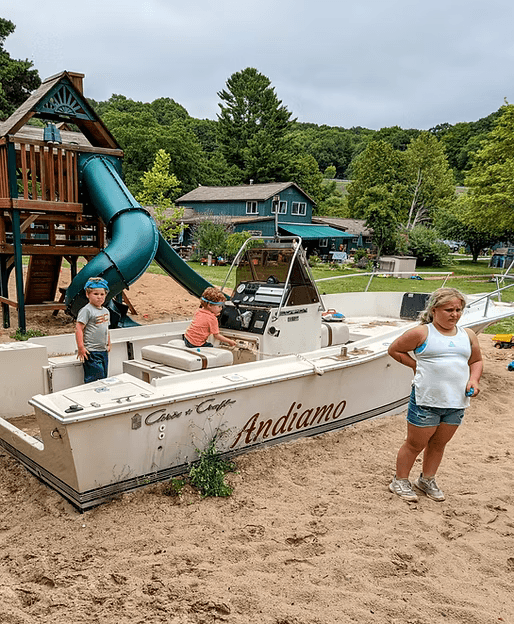 Children play around a boat nestled in sand near a playground with a slide.