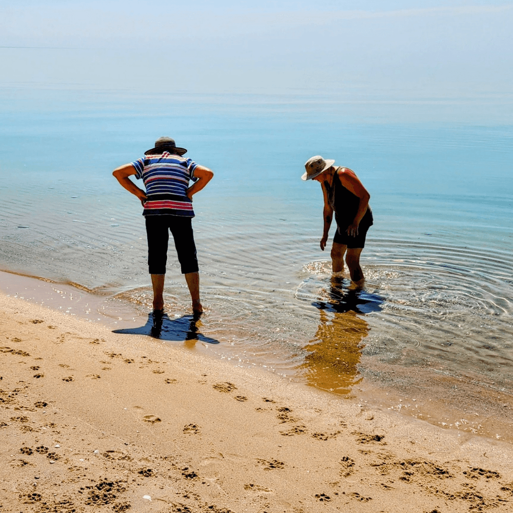 Two people stand at the edge of a calm beach, inspecting the shallow water.