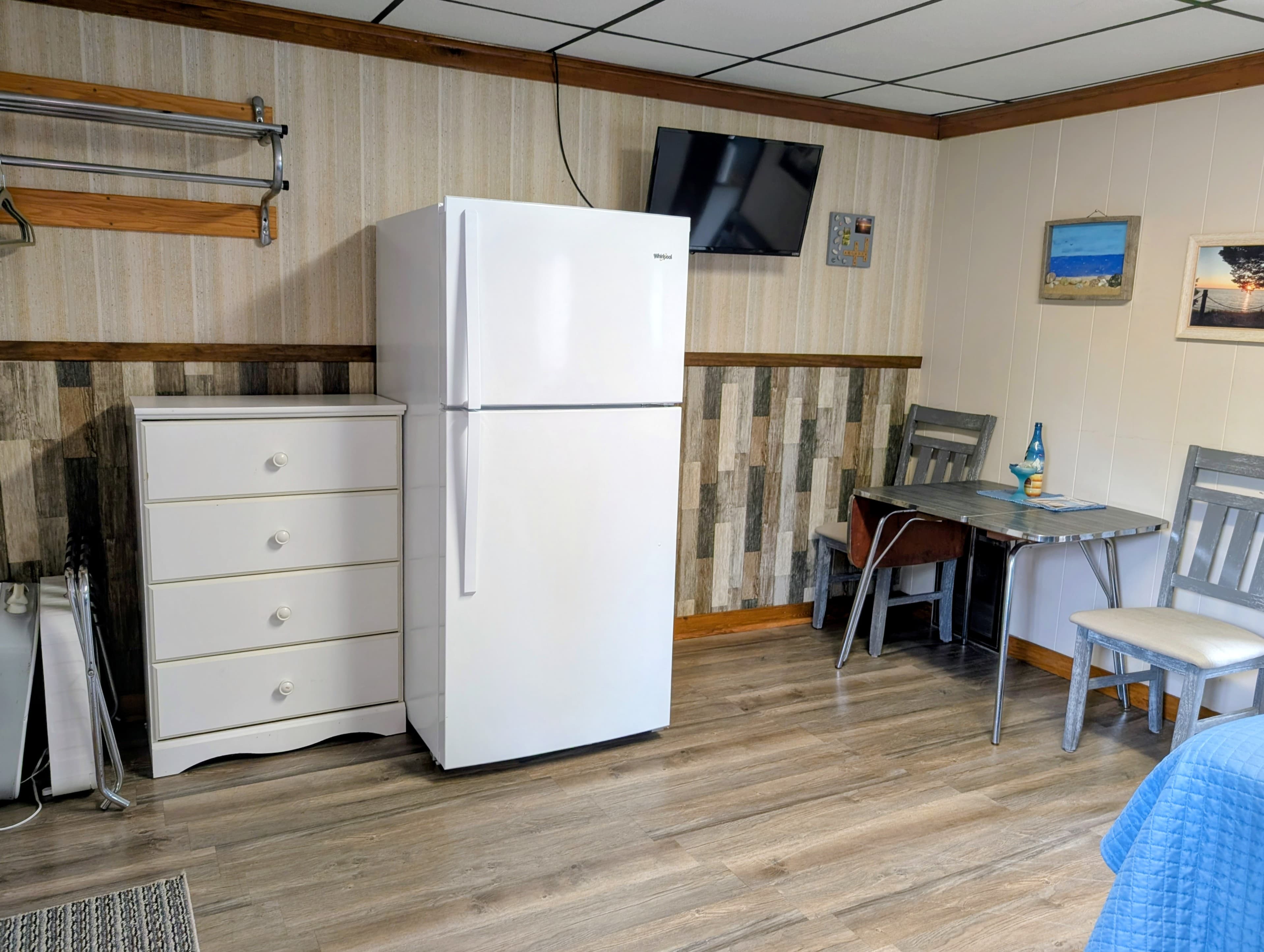 A compact kitchen area featuring a white refrigerator, a dress drawer, a small dining table with chairs, and a wall-mounted TV.