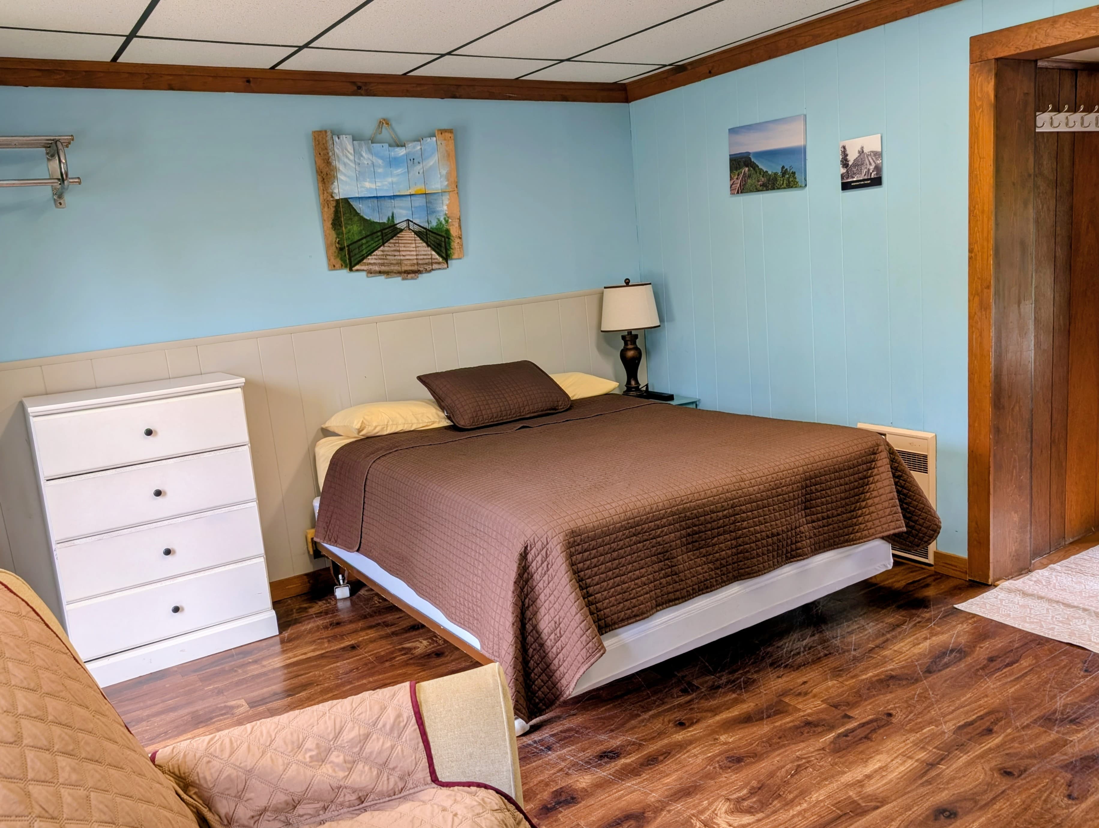 A cozy bedroom featuring a brown quilt, white furniture, and light blue walls adorned with artwork.