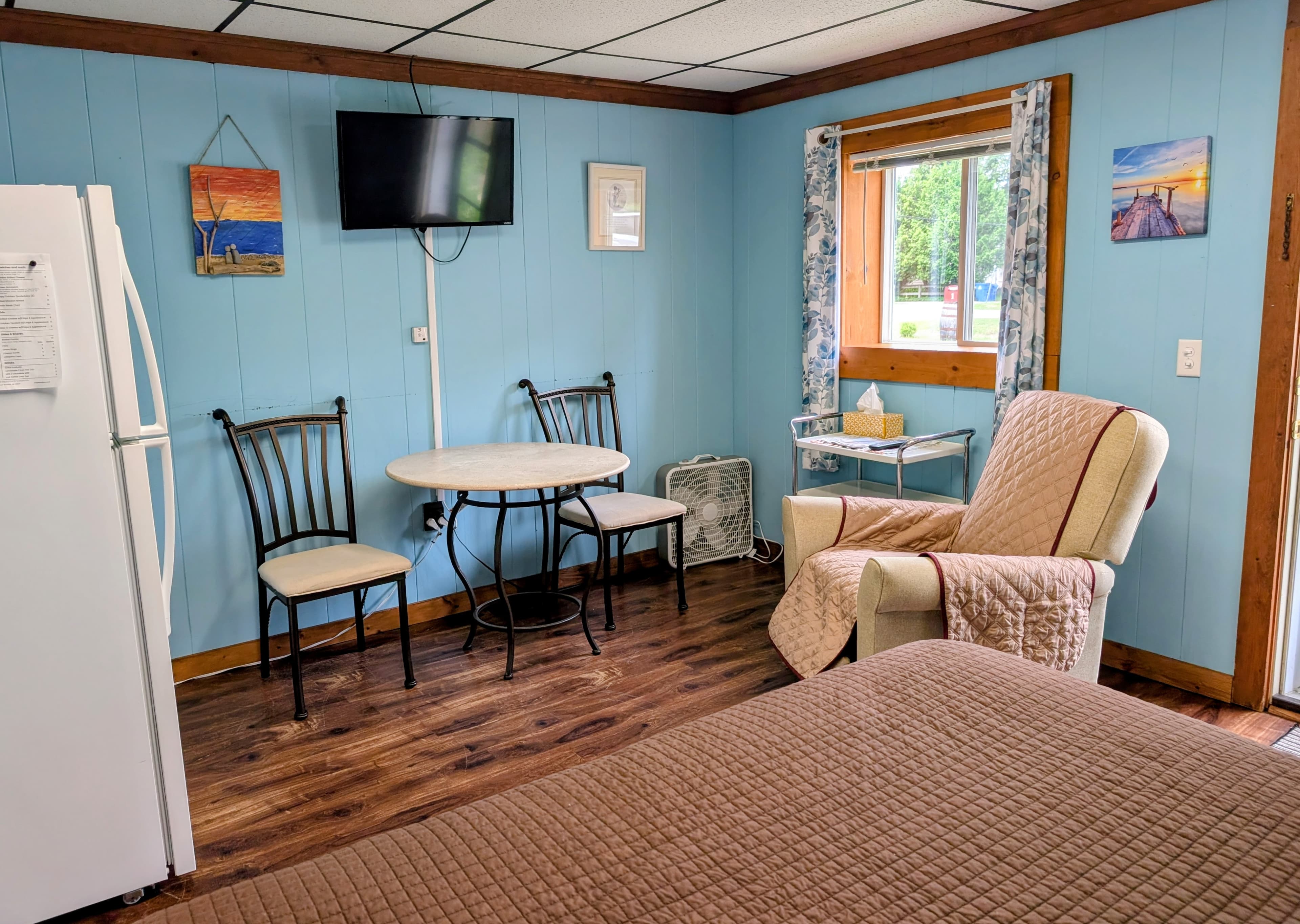 A cozy room featuring a bed, a small dining table with chairs, a recliner, a refrigerator, a TV, and light blue walls.