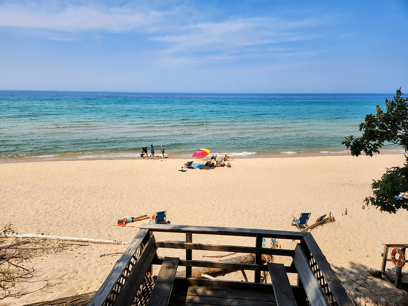 A view from a wooden deck overlooking a sandy beach with people, colorful umbrellas, and calm blue waters.