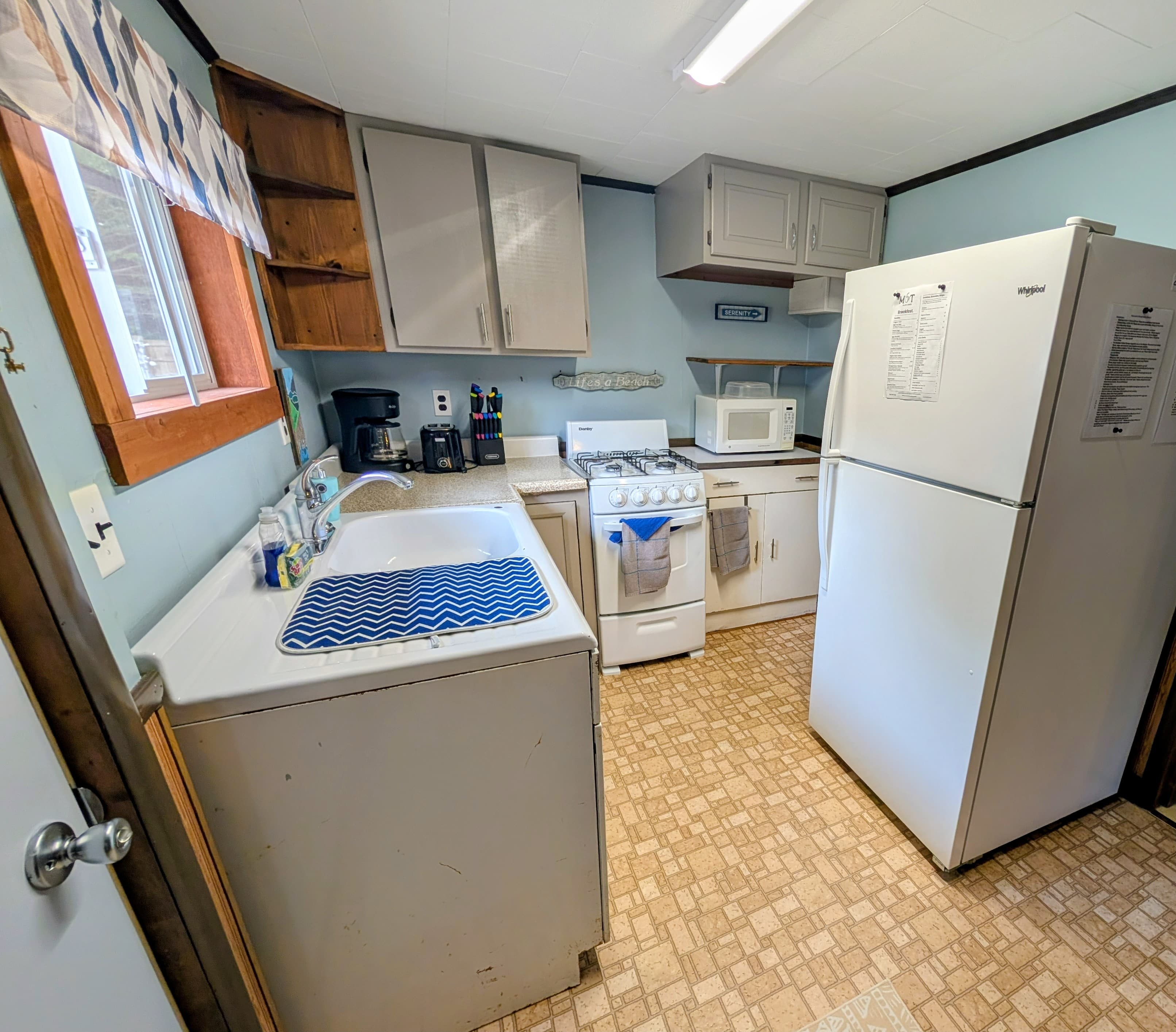 A small, functional kitchen featuring a sink, stove, microwave, and refrigerator with a light blue wall and linoleum flooring.