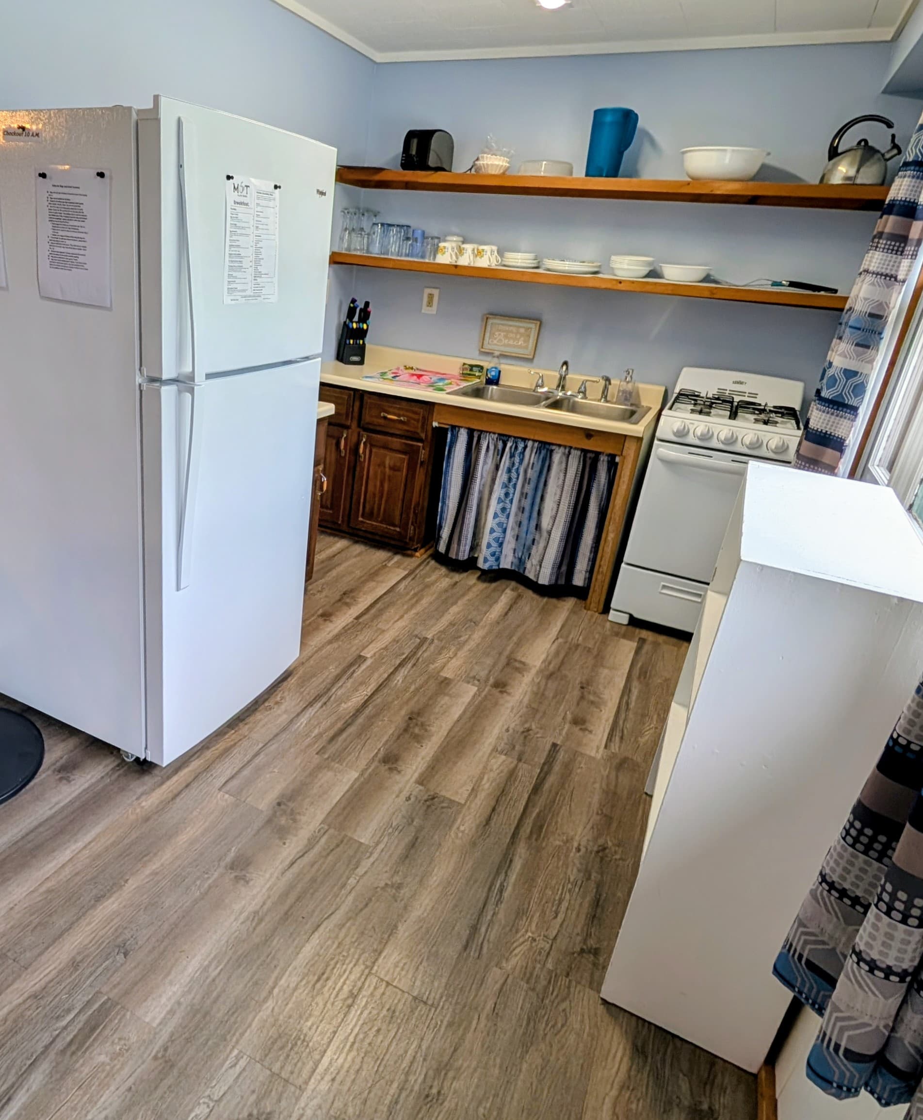 A compact kitchen featuring a refrigerator, stove, sink, and open shelving.
