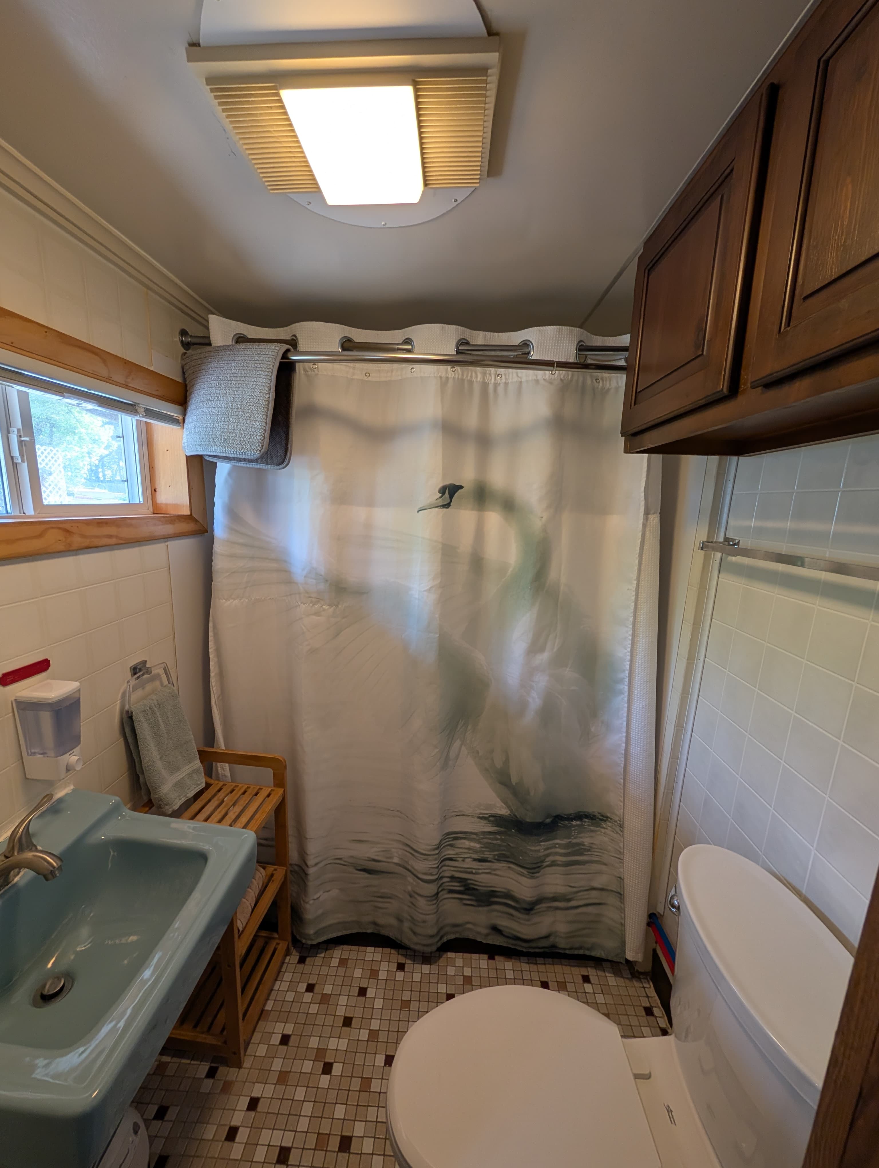 A small bathroom featuring a blue sink, white toilet, wooden shelving, and a shower curtain with a swan design.