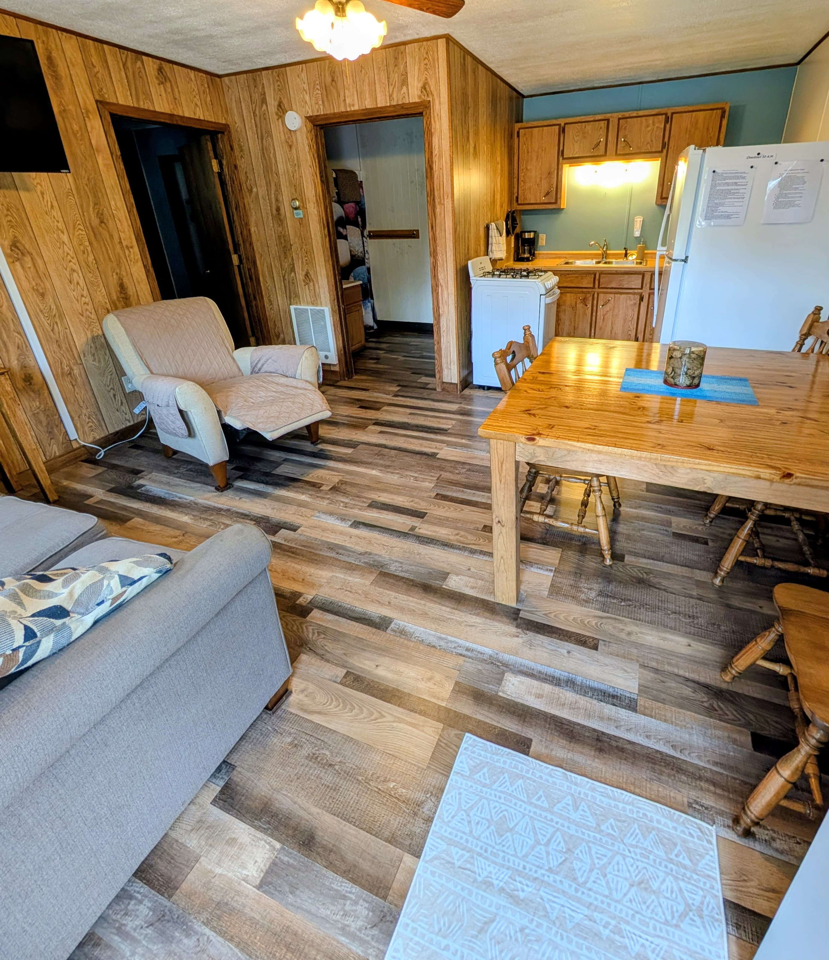 A cozy living area with wooden flooring, a comfortable chair, a dining table, and a kitchen visible in the background.
