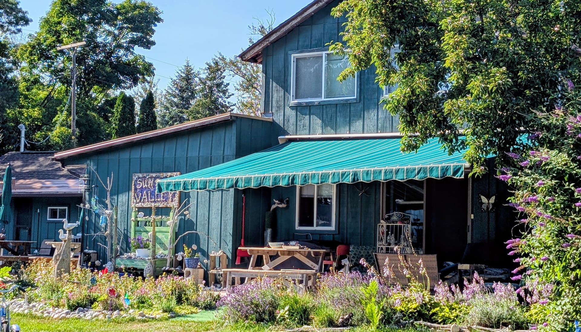 A two-story green house with a teal awning and a colorful garden in front.