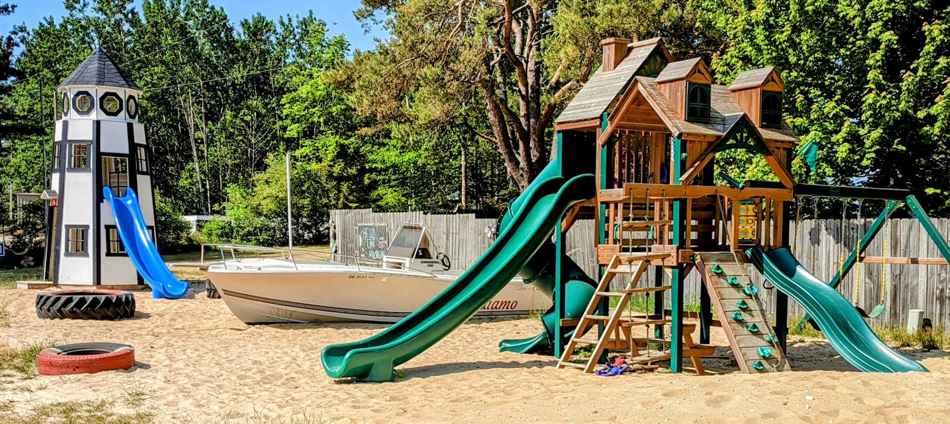 A playground featuring a wooden play structure with slides, a lighthouse-shaped slide tower, and a boat in a sandy area.