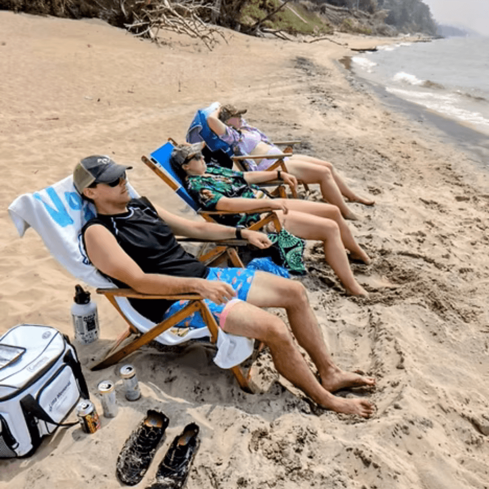 Four people relax on beach chairs along a sandy shore, enjoying drinks by the water.